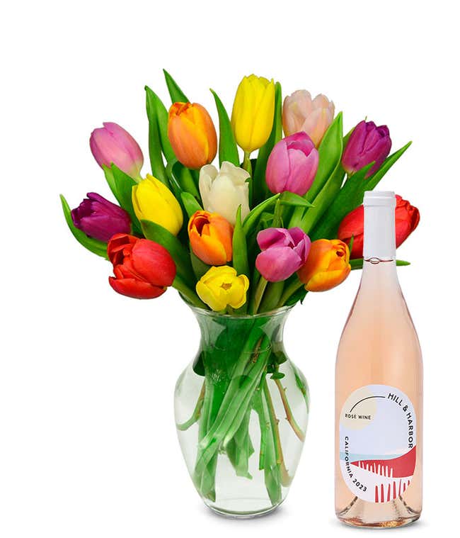 A vibrant array of tulips in red, pink, yellow, orange, and white, each bloom vivid and fresh, stands in a clear glass vase. Accompanying the flowers is a bottle of rose wine with a minimalist pink label, indicating a 2023 vintage from California.
