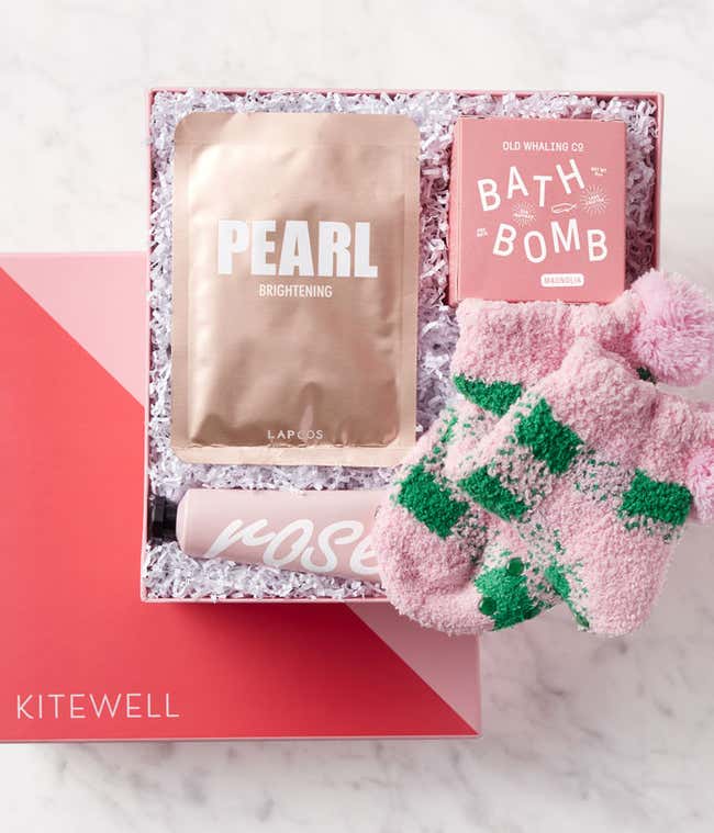 Gift box with a gold pearl brightening face mask, pink magnolia-scented bath bomb, rose-scented hand cream, and a pair of fuzzy pink and green plaid socks with pom-poms, all arranged on white crinkle paper in a pink box.