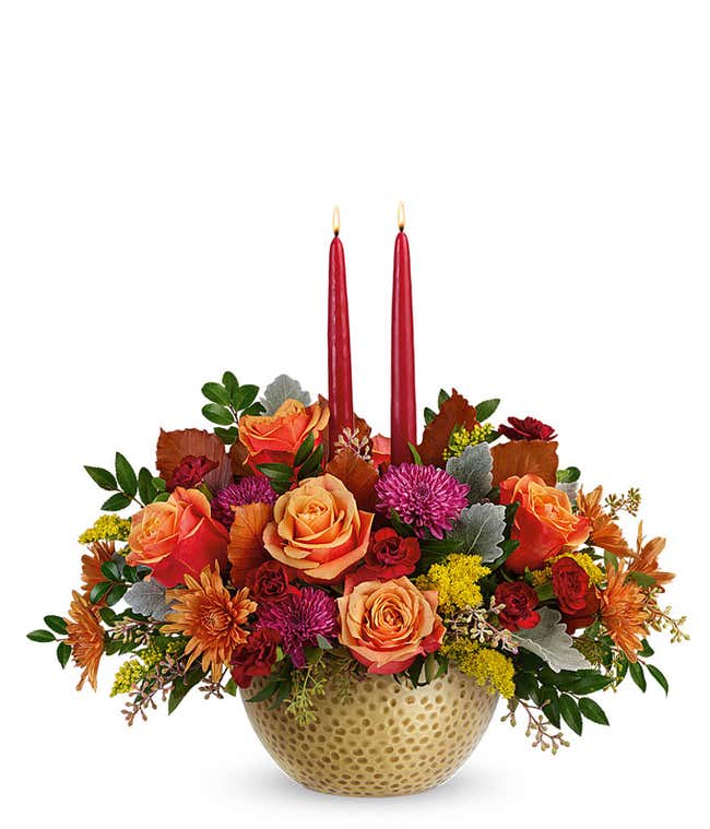 A rich floral arrangement featuring orange roses, maroon miniature carnations, rust and purple cushion spray chrysanthemums, solidago, brown copper beech, dusty miller, seeded eucalyptus, and huckleberry, complemented by burgundy candles, all beautifully 