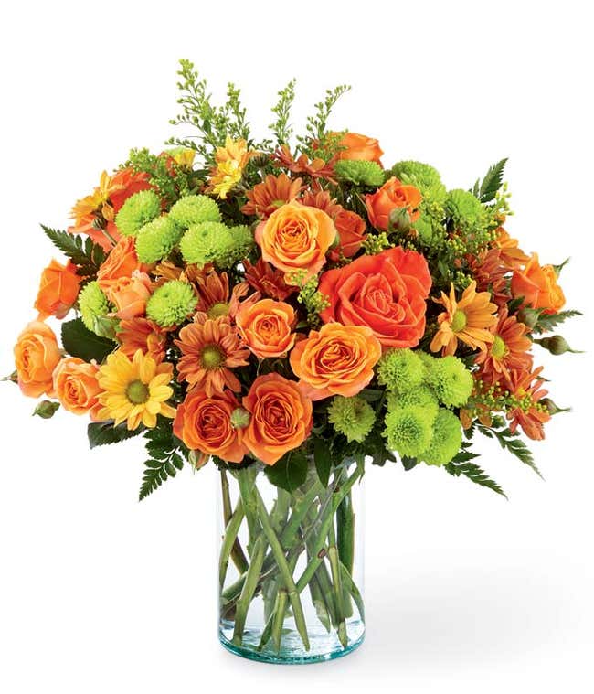 A vibrant floral arrangement featuring orange roses, orange spray roses, butterscotch daisies, green button poms, and solidago, all beautifully displayed in a clear glass vase.