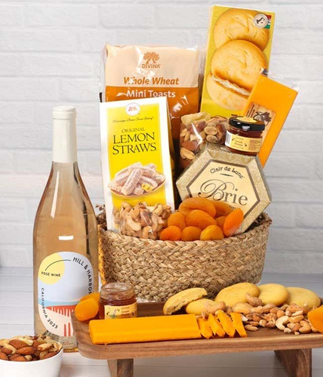 A two-toned woven basket filled with two cheeses, lemon flavored baked goods, crackers, dried apricots, gourmet nuts, and honey, paired with a bottle of ros&eacute; wine.