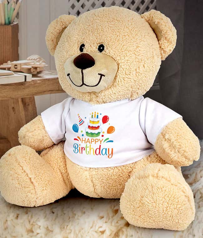 Plush tan teddy bear wearing a white t-shirt printed with a colorful birthday cake, balloons, and the words Happy Birthday, shown seated against a white background.